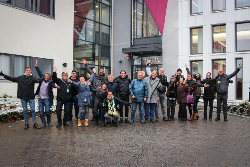 Todos os participantes do EMBRACE (Finlândia, Portugal, Brasil e Colômbia) em pé e agachados, acenando para a foto na fachada da HAMK University of Applied Science em, Häamenlinna, Finlândia.