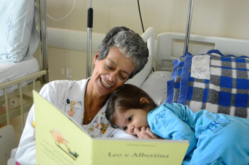 Voluntário com livro conta história para criança internada em hospital. Criança está com a cabeça no ombro da voluntária e olha atenta para o livro.