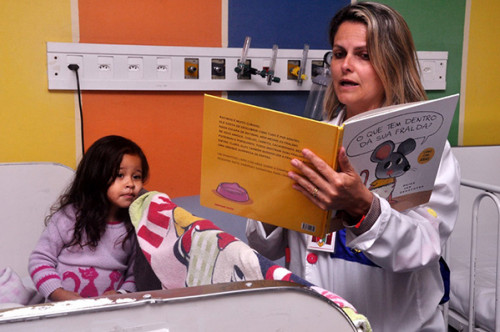 Voluntário com livro na mão conta história para criança internada em hospital. Criança olha atenta.